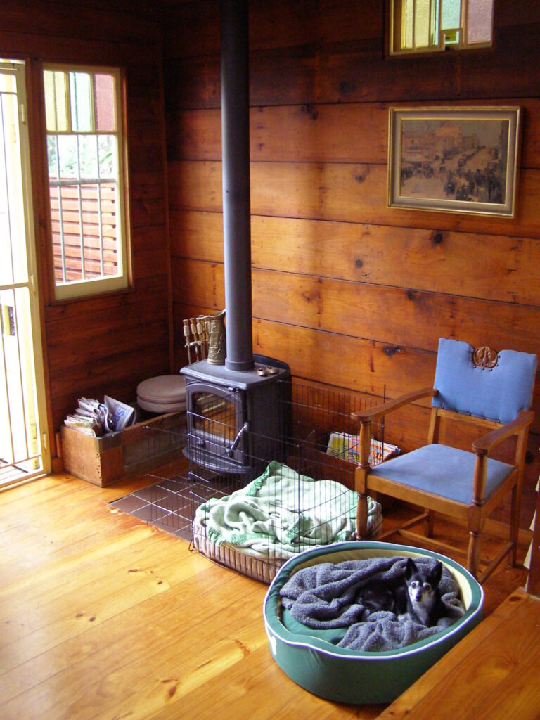 A small black and white toy dog resting in a dog bed beside a wood stove in a wood panelled room.